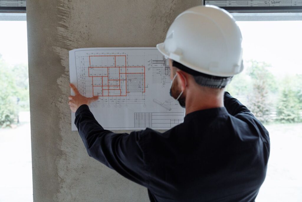 مهندس يرتدي خوذة ويفحص مخططًا معماريًا بجوار نافذة في مبنى قيد العمل